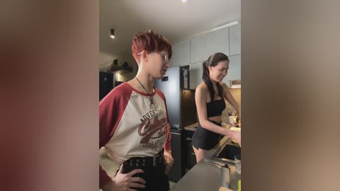Video of two young women in a modern kitchen. One with short red hair in a long-sleeve shirt, the other with long brown hair in a sports bra and shorts, both looking at a stove.