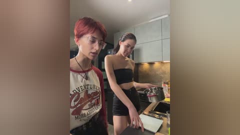 Video of two women in a modern kitchen; one with short red hair, wearing a \"Coca-Cola\" T-shirt, the other with long brown hair, dressed in a black bandeau top and shorts.