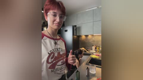 Video of a young person with short red hair, wearing glasses and a \"Adventure Club\" t-shirt, giving a thumbs-up in a modern kitchen with stainless steel appliances.