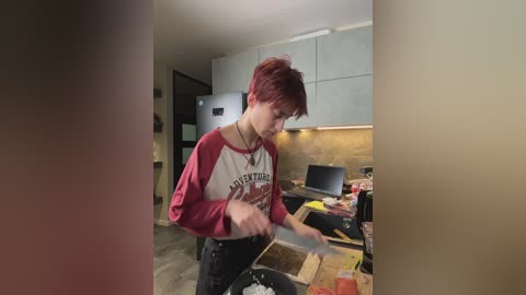 Video of a young person with short red hair, wearing a red and white long-sleeve shirt, standing in a modern kitchen with white cabinets, countertops cluttered with items, and a black fridge.