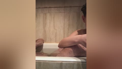 A video of a man's lower torso in a beige-tiled bathroom, sitting in a bathtub with his legs crossed, his erect penis visible.