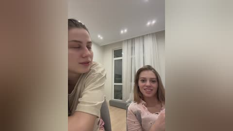 Media: Video of two young women sitting in a modern living room. One with light skin, long hair, beige shirt, and closed eyes; the other with light skin, long hair, pink shirt, and smiling.