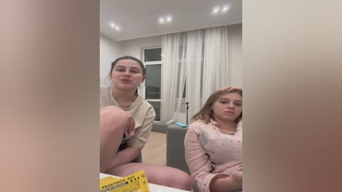 Media: Video of two young women, one with light brown hair and a beige top, the other with blonde hair and a pink top, sitting on a gray couch in a modern living room.