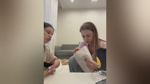 Media: Video of a woman with light skin and straight brown hair, wearing a pink top, reading a book at a white table in a minimalistic room with a gray sofa in the background.