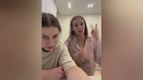 Media: Video of two young women in a modern, well-lit room. One with fair skin, dark hair, and a beige top, and another with fair skin, light blonde hair, and a pink blouse.