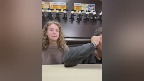 Media: Video of a young, curly-haired girl in a gray sweater, seated at a table in a restaurant with a dark wooden backdrop and a group of toy figures in uniforms.