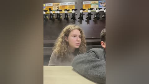 Media: A video of a young woman with curly blonde hair, wearing a grey sweater, seated in a dark, cafeteria-like room with multiple mic stands and a yellow menu board in the background.
