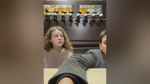 Media: Video of a young woman with curly hair in a beige sweater, seated next to a man in a gray jacket, in a dimly lit room with black uniforms and hats displayed on a wall.