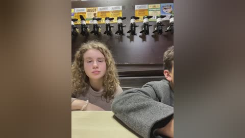 Media: Video of a young girl with curly blonde hair in a gray top, sitting at a table in a classroom with rows of empty chairs, blackboard, and posters in the background.