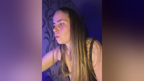 Media: Video of a young Caucasian woman with long, straight brown hair, wearing a black lace bralette, illuminated by purple and blue lighting against a dark, patterned wallpaper.
