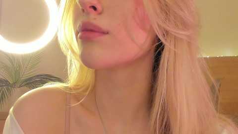 Media: Video of a fair-skinned woman with long, blonde hair, wearing a white off-shoulder top, partially illuminated by a ring light.