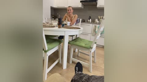 Media: Video of a smiling woman with blonde hair, wearing a white tank top, seated at a white table in a modern kitchen.