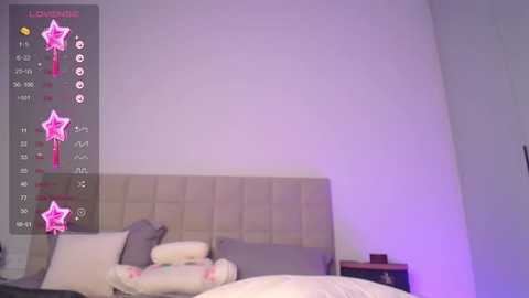 Media: Video of a bedroom with a white, tufted headboard bed, light gray pillows, and a purple nightstand lamp. A smartphone app interface overlay shows a pink star and stats.