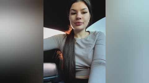 Media: Video of a young woman with long brown hair, wearing a light grey long-sleeve top, seated in a car, slightly out of focus, with a dark background.