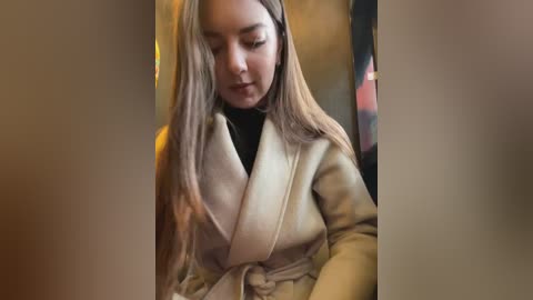 Media: A video of a young woman with long, straight blonde hair, wearing a beige coat and black turtleneck, looking down in a dimly lit, indoor setting.