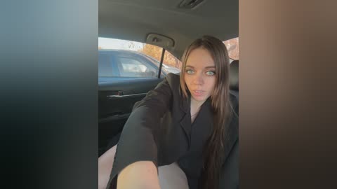 Media: Video of a young woman with long brown hair, wearing a black top, sitting in a car, looking directly at the camera.