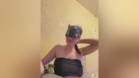 Video of a young woman with light skin, wearing a black Batman mask and black strapless top, seated against a beige wall with a patterned bedspread in the background.