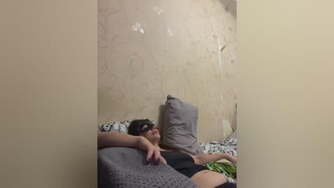 Video of a woman with fair skin, wearing a black mask and black shirt, lying on a bed with patterned bedding, against a beige wallpapered wall.