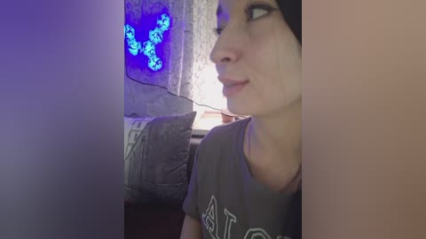 Video of a light-skinned woman with dark hair, wearing a grey T-shirt, looking to the side in a dimly lit room with a blue glowing LED wall panel.