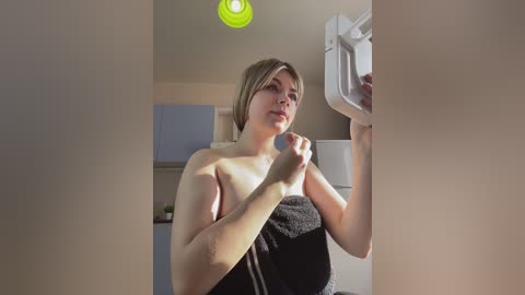 Video of a topless, fair-skinned woman with short blonde hair, standing in a modern kitchen, holding a white dish rack. She wears a black towel and looks contemplative.