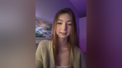 Media: Video of a young Caucasian woman with long brown hair, light skin, and a slim build, smiling. She wears a beige knitted cardigan over a white tank top. Background features a colorful abstract painting and purple lighting.