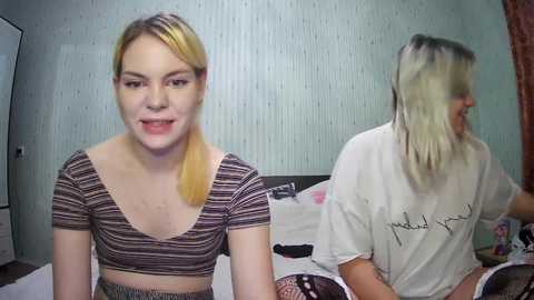 Media: Video of two young women in a bedroom: one with blonde hair, wearing a striped crop top, and the other with dyed hair, wearing a white t-shirt.