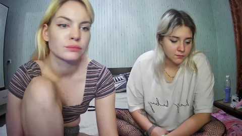 Media: Video of two young women with light skin and blonde hair, sitting on a bed in a bedroom with light green wallpaper. One wears a striped top, the other a loose white shirt.