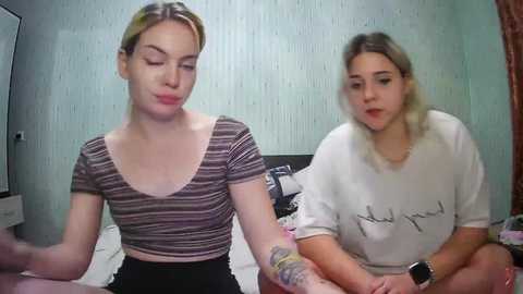 Media: Video of two women with fair skin, one with blonde hair in a striped top, the other with brown hair in a white shirt, sitting on a bed in a dimly lit room.