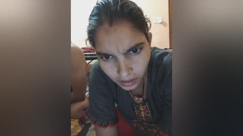 Video of a young woman with medium brown skin and dark hair pulled back, wearing a black t-shirt, leaning over a child on a bed in a dimly lit room.