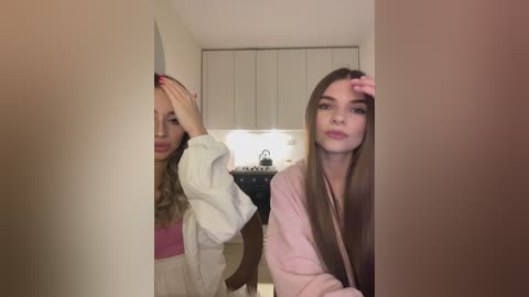 Media: Video of two young women, one with blonde hair, the other brunette, standing side by side in a modern kitchen. The blonde woman, wearing a white top, has her hand on her head, while the brunette, in a pink robe, looks directly at the camera.