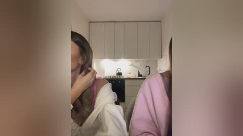 Media: Video of a woman in a white bathrobe, hair disheveled, standing in a minimalist, modern kitchen with white cabinets, a white marble backsplash, and stainless steel appliances.