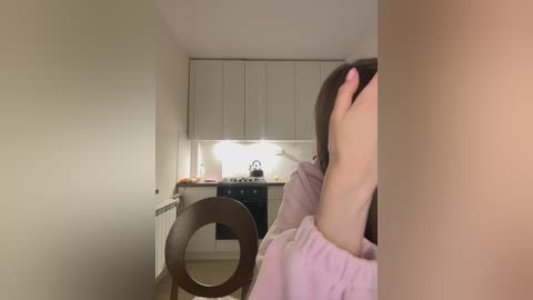 Media: Video of a small, dimly lit kitchen with a woman in a light pink top sitting at a table, covering her face with her hand.