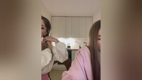 Media: Video of a woman with long brown hair, wearing a white robe, applying makeup in a modern, dimly lit kitchen with white cabinets and a marble backsplash.