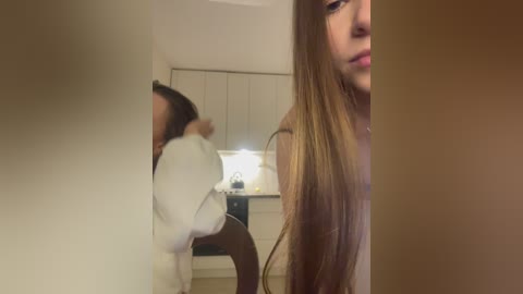 Media: Video of a young woman with long, straight, light brown hair, wearing a white sweater, standing in a dimly lit kitchen with beige cabinets and a counter. Her face is partially obscured by a vertical object in the foreground.