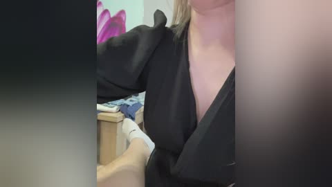 A video showing a close-up of a woman with light skin, wearing a black dress with a deep V-neckline. Her blonde hair is partially visible. The background includes a bed with blue sheets and a pink pillow.