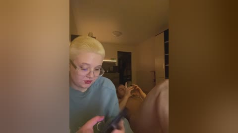 A video of a fair-skinned, blonde-haired woman with glasses, wearing a blue shirt, looking at a smartphone in a dimly lit room with beige walls and a kitchen visible in the background.