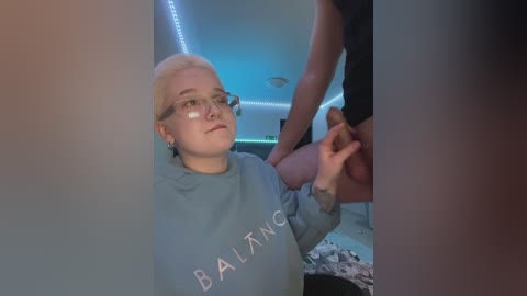 Video of a young, fair-skinned woman with short platinum blonde hair, wearing glasses and a \"Balancing\" sweatshirt, sitting in a dimly lit, modern room with blue LED strip lights on the ceiling. Another person, partially visible, is standing behind her.