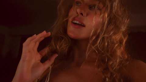 Media: Video of a young, fair-skinned woman with curly, shoulder-length blonde hair, wearing a top, playfully touching her hair with her right hand. The background is dimly lit, creating a warm, intimate atmosphere.