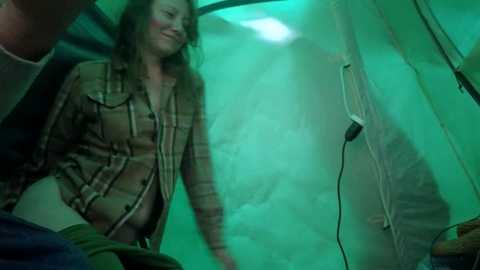Media: A video captures a grinning woman with long, wavy hair, wearing a plaid shirt, inside a green-lit tent. She's holding a black cord, with camping gear visible in the background.