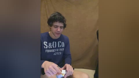 Video of a young man with curly hair, wearing a navy blue long-sleeved shirt with \"Sailor Found in 1968\" text, seated in a beige room, holding a small object.