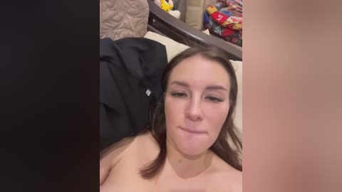 A candid video of a topless woman with fair skin and brown hair, smiling, lying on a bed with a black sheet, surrounded by colorful snacks on a table in the background.