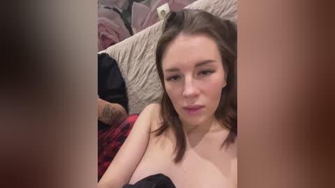 Media: A video captures a young, fair-skinned woman with shoulder-length brown hair, lying on a bed with a black and red checkered blanket. She has a tattoo on her forearm and looks contemplative.