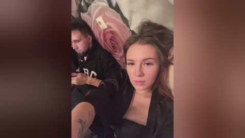 Media: A video of two people, a man and a woman, sitting on a bed with a pink rose and a white pillow. The woman, with brown hair, wears a black top, and the man, with a beard, wears a black hoodie.