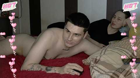 Video of a nude man with tattoos lying on a red blanket, while a woman with dark hair and a black shirt rests beside him, in a dimly lit room.