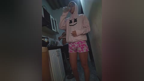 Video of a young woman with light skin, wearing a pink hoodie with a smiley face and pink shorts with a floral pattern, standing in a narrow, dimly lit hallway.