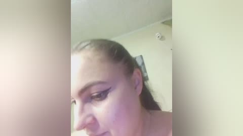 A close-up video of a woman with light skin, dark hair pulled back, wearing black eyeliner, and a white top, taken in a dimly lit room with beige walls and a ceiling light.
