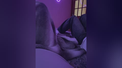 Media: Video of a partially visible person with a shaved head and exposed buttocks lying on a bed with purple lighting. The background shows a wooden screen and a soft, patterned blanket.