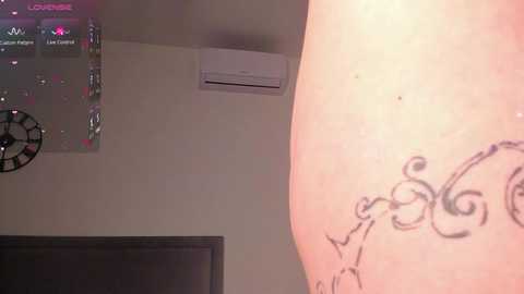 Video of a close-up of a person's lower back showing a black, elegant, swirling tattoo design. Background features a beige wall with an air conditioner, a clock, and a television partially visible.