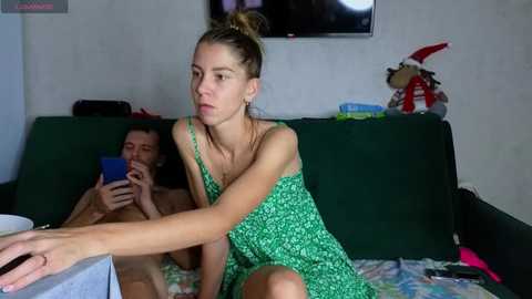 Video of a young woman in a green patterned dress, sitting on a couch, playing a video game with a man in the background.