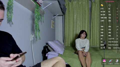 Media: Video of a woman sitting on a bed in a dimly lit room, wearing a white knit sweater and shorts, while a man with a smartphone stands nearby.